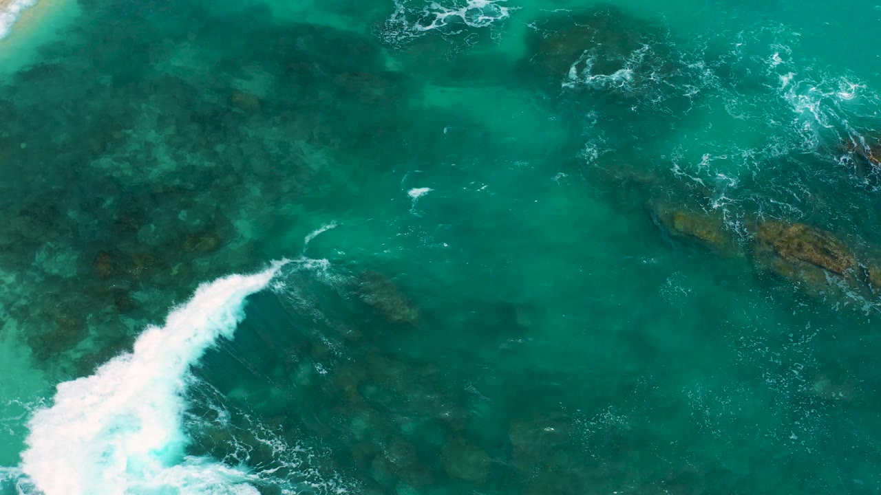 A larger white wave plume crosses the lower left of the overhead view at Secrets Marquis Los Cabos, Mexico, with dark rocks visible beneath teal water, no visible people.