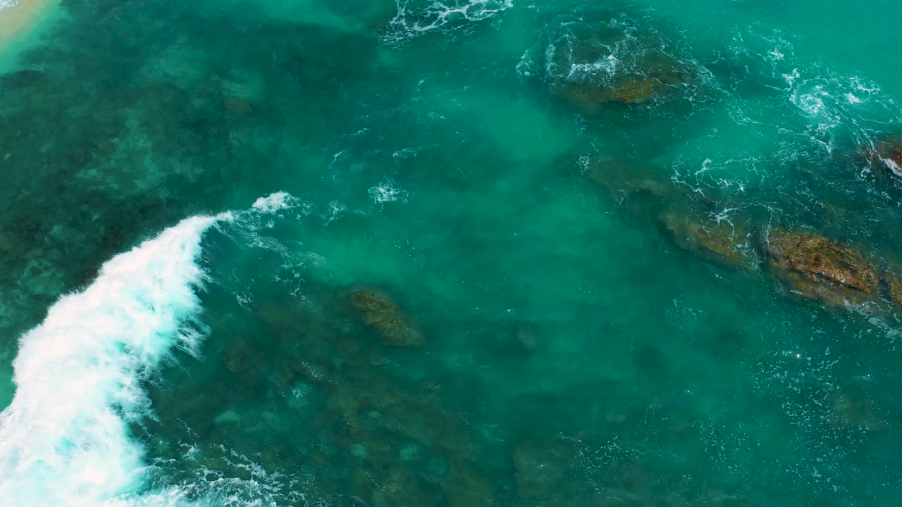 Overhead view from Secrets Marquis Los Cabos, Mexico, with white surf pushing diagonally through turquoise water and darker rock shapes below the surface, no visible people.