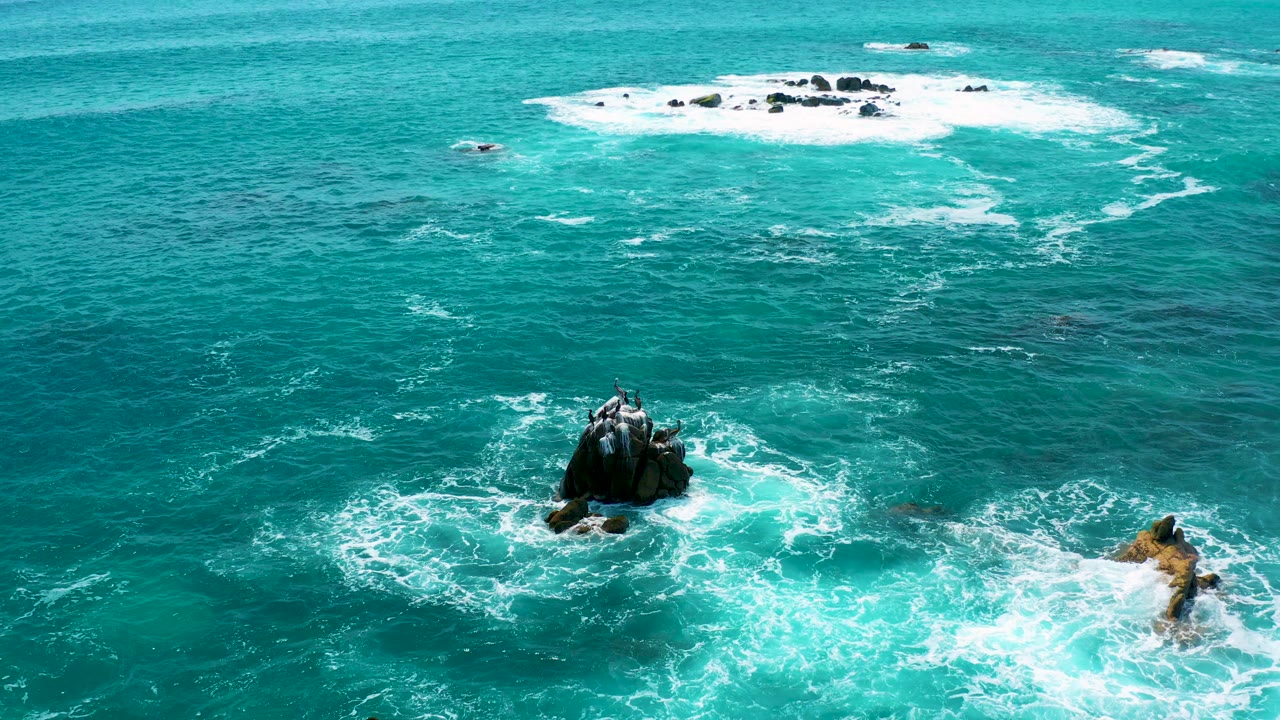Aerial frame from Secrets Marquis Los Cabos, Mexico, showing turquoise water, a dark rock in the lower left, tan rocks to the lower right, distant white reef wash, and no visible people.