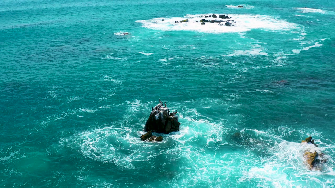 Aerial frame from Secrets Marquis Los Cabos, Mexico, showing white surf circling a dark central rock, smaller tan rocks at the right edge, broad teal water, and no visible people.