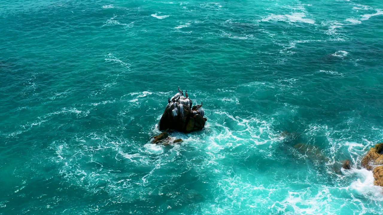 Aerial frame from Secrets Marquis Los Cabos, Mexico showing a dark rock centered in turquoise ocean with white foam forming around its base and no visible people.