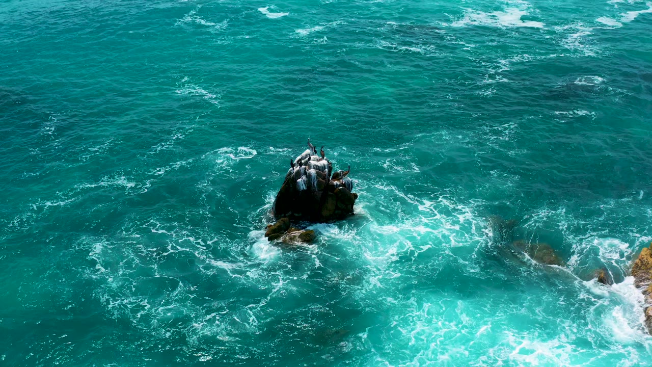 Aerial shot over Secrets Marquis Los Cabos, Mexico showing the dark rock above churning blue water, with foam trailing toward the right edge and no visible people.