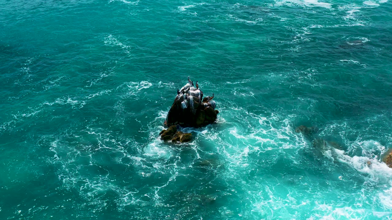 Aerial frame from Secrets Marquis Los Cabos, Mexico where swirling white foam circles the central rock against teal water, with no visible people.