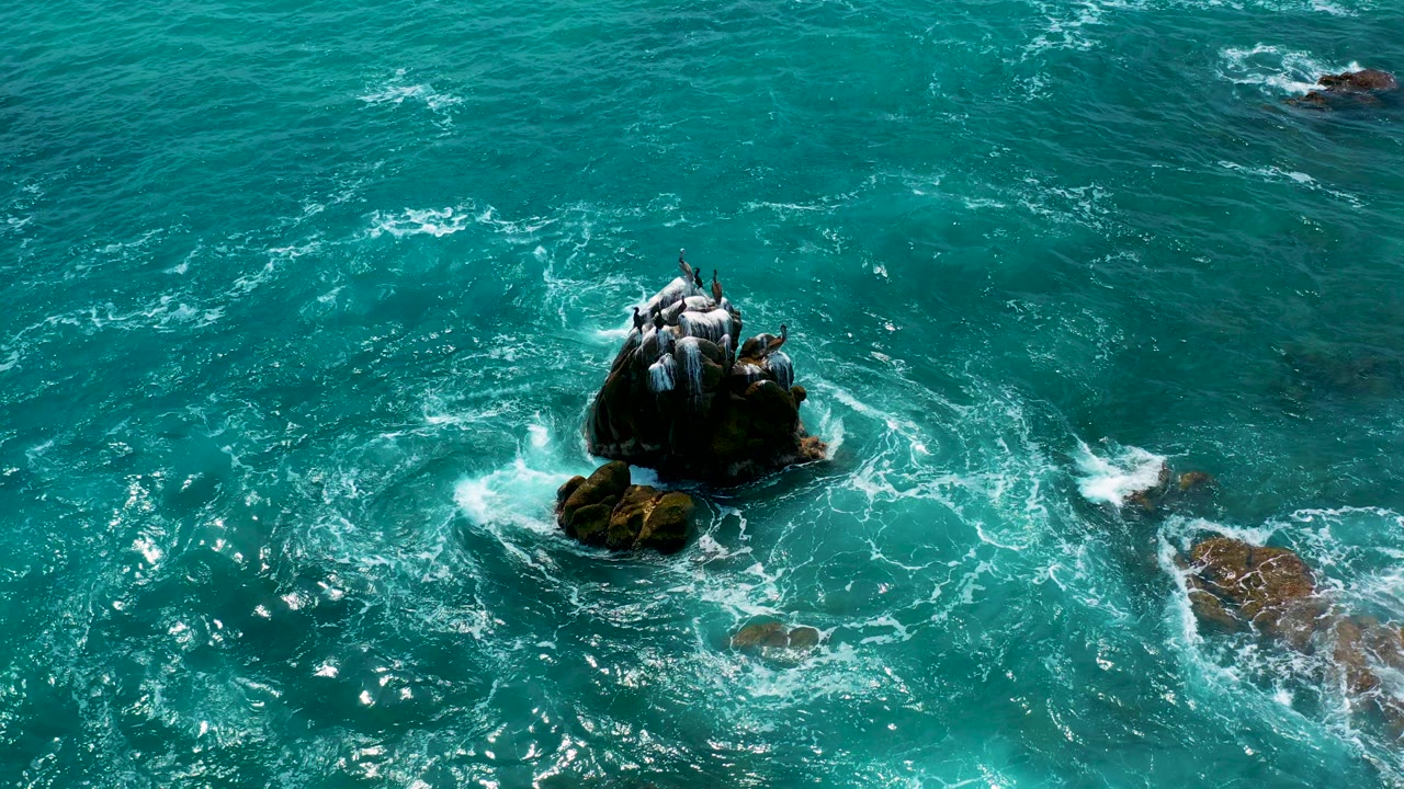 Overhead view at Secrets Marquis Los Cabos, Mexico, showing turquoise water and white foam circling a dark rock stack with seabirds perched above it and no visible people.