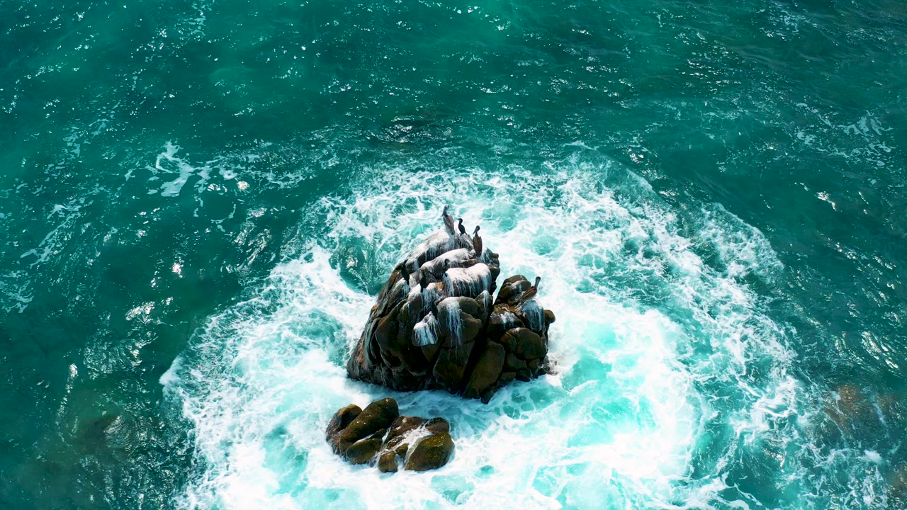 Top-down aerial frame at Secrets Marquis Los Cabos, Mexico, showing teal water and white foam surrounding a compact cluster of dark rocks, with no visible people.