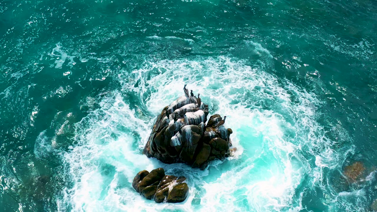 Top-down drone-style frame at Secrets Marquis Los Cabos, Mexico, showing dark boulders encircled by breaking white water and darker open sea above, with no visible people.