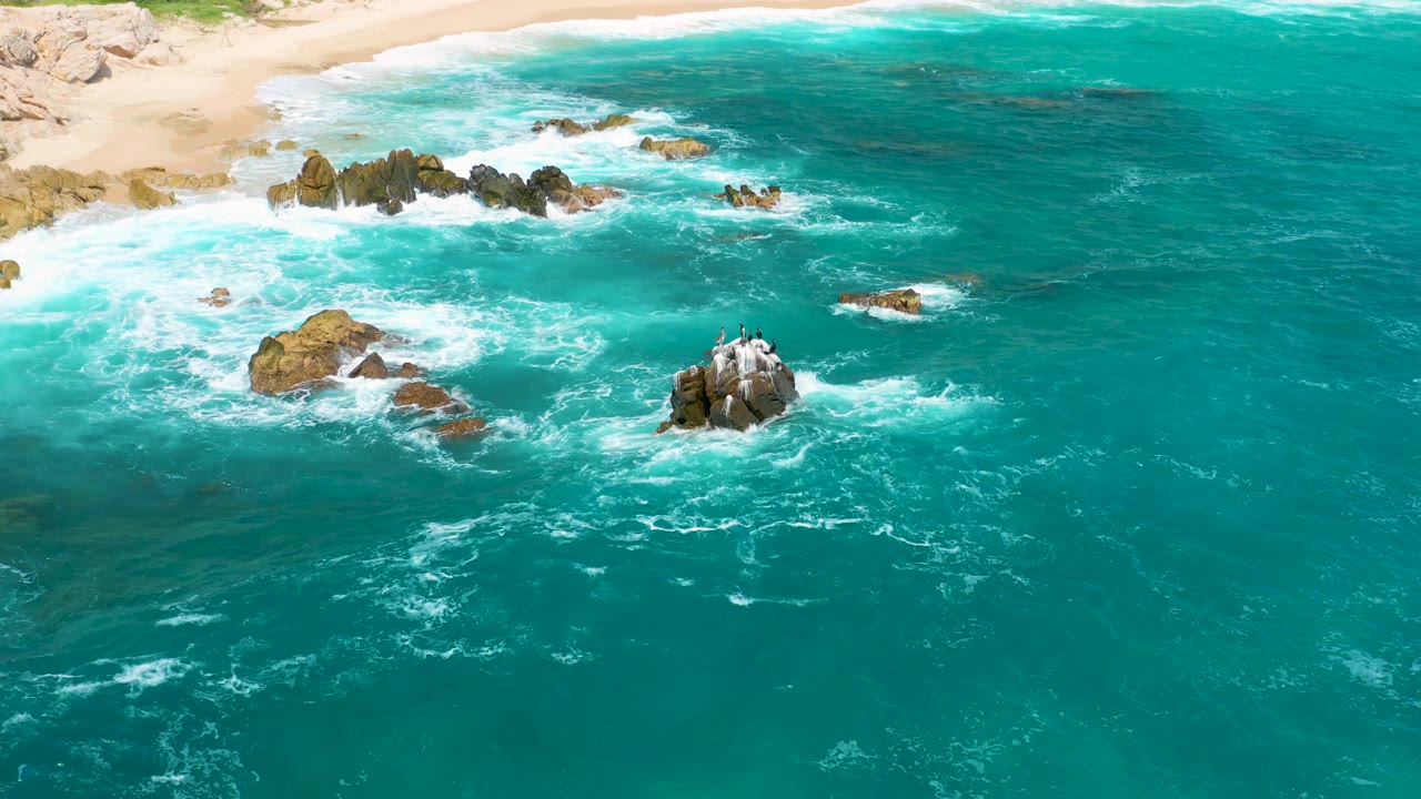 Aerial view at Secrets Marquis Los Cabos, Mexico, with blue-green ocean filling the frame, foam breaking across several rocks, and no visible people.