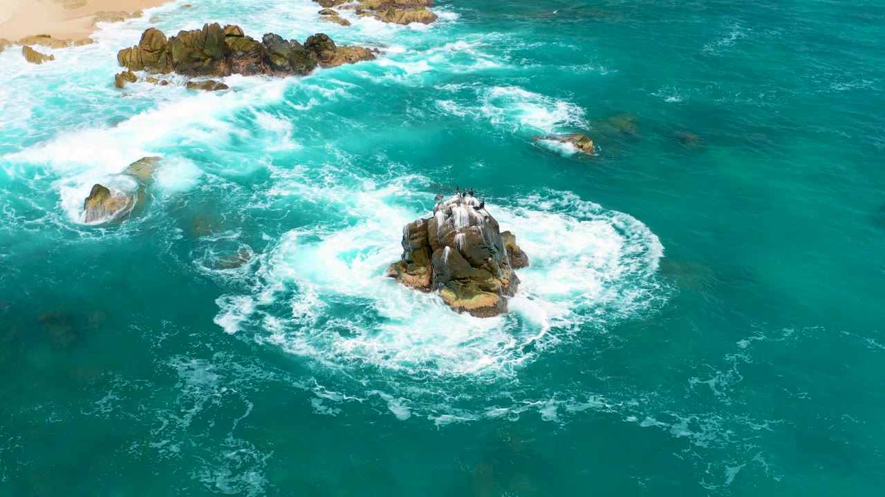 Aerial frame from Secrets Marquis Los Cabos, Mexico showing turquoise ocean, scattered rocky outcrops, sandy shore near the top edge, and no visible people.