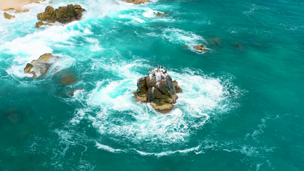 Aerial frame from Secrets Marquis Los Cabos, Mexico showing a larger ring of white foam around the central rock, with open turquoise water to the right and no visible people.