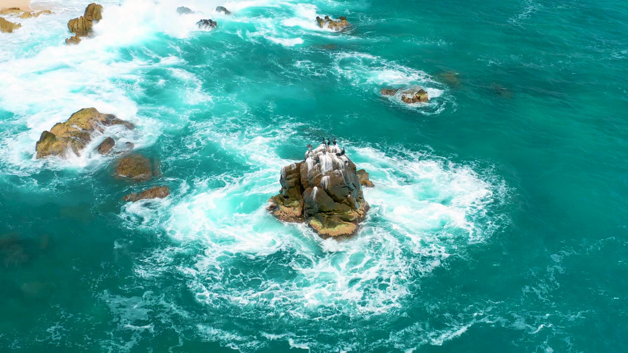 Aerial view at 0:03 over Secrets Marquis Los Cabos, Mexico, with the central rock lower in frame, breaking white waves to the left, and no visible people.