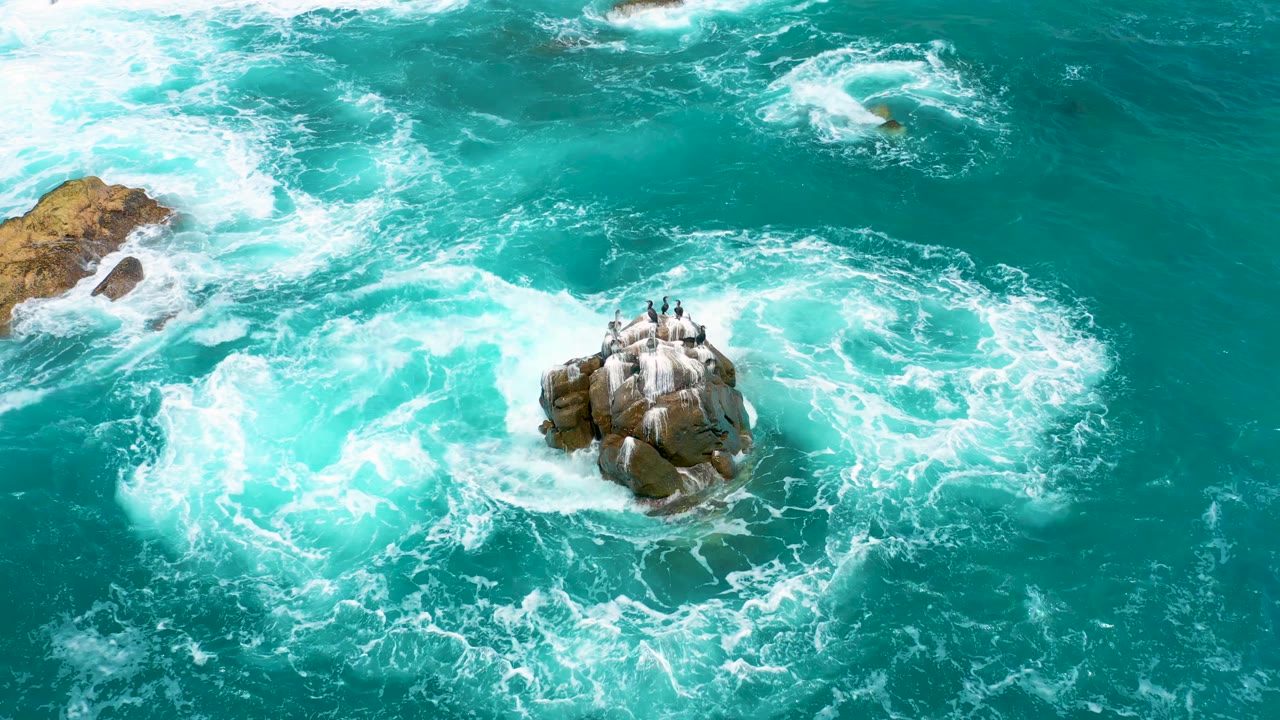 White foam spreads wider around the rocky island in a high aerial view of the Secrets Marquis Los Cabos coastline, with no visible people.