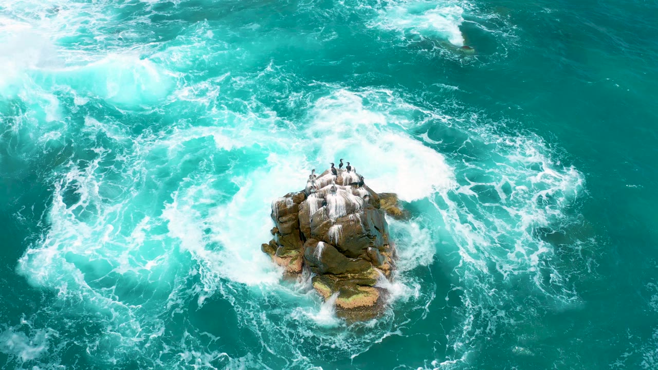 An overhead frame shows the rock island lower in the image while broad white foam patches move through turquoise water at Secrets Marquis Los Cabos, with no visible people.