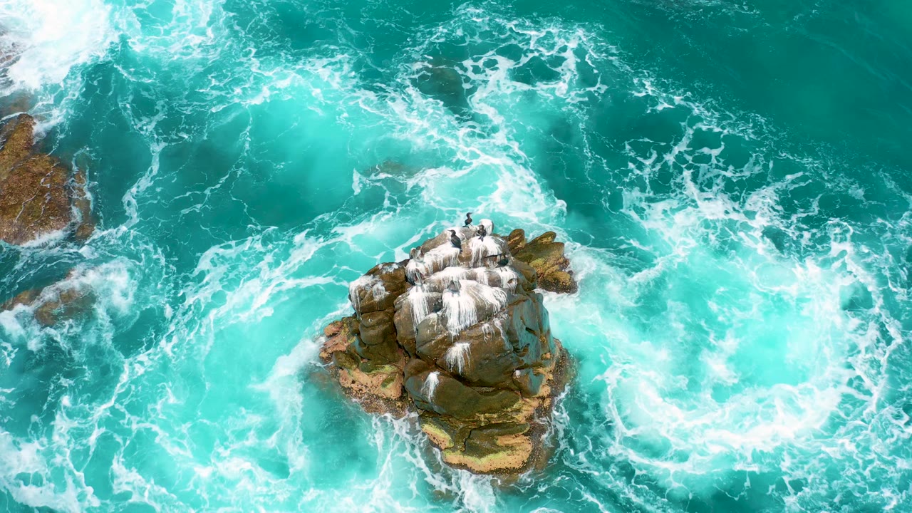 Overhead frame from Secrets Marquis Los Cabos, Mexico, where swirling foam curls to the left of the rock formation and clear teal water fills the upper frame, no visible people.