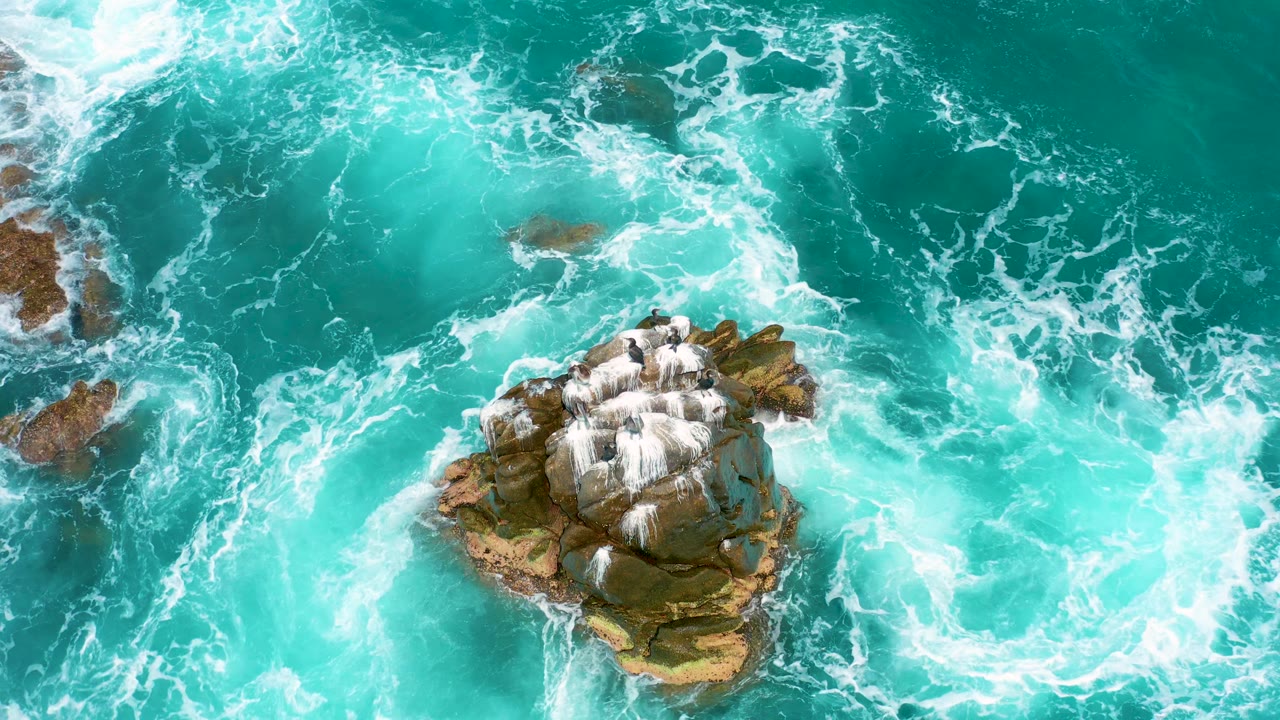 Top-down Secrets Marquis Los Cabos, Mexico view with the rock formation lower in frame as white foam trails through bright turquoise water, no visible people.