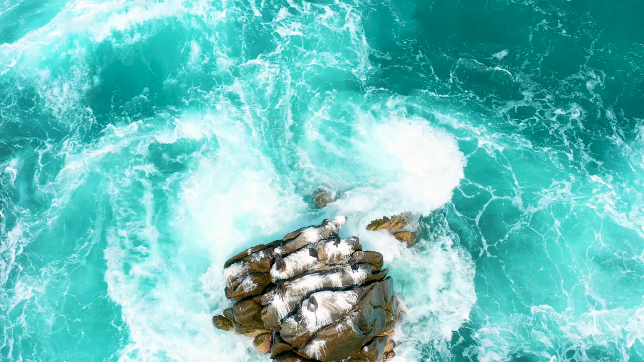 Aerial frame of clear blue-green water curling into whitewater beside a rocky outcrop at Secrets Marquis Los Cabos, Mexico, with no visible people.