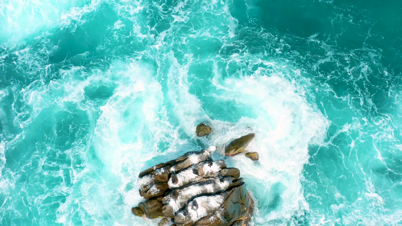 Top-down view of foamy surf swirling near exposed rocks in bright turquoise ocean at Secrets Marquis Los Cabos, Mexico, with no visible people.