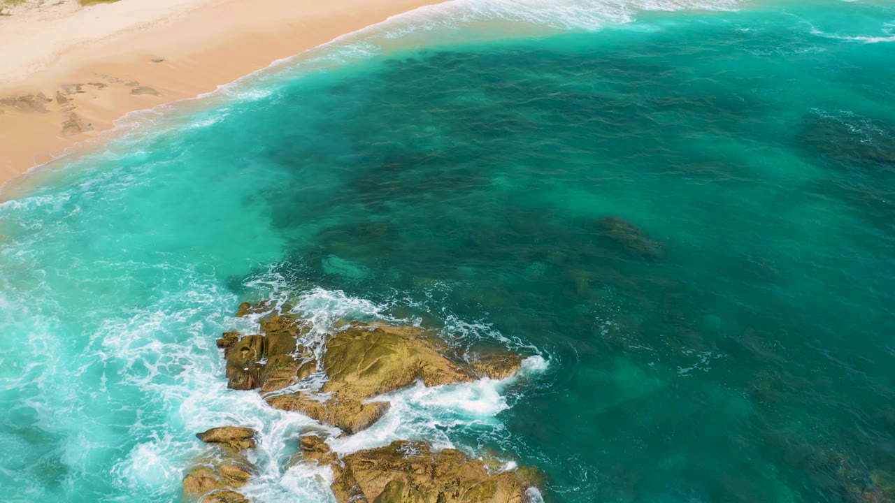 Aerial frame at Secrets Marquis Los Cabos, Mexico with the rocks lower in the image, turquoise water across the center, beach along the top, active foam, and no visible people.
