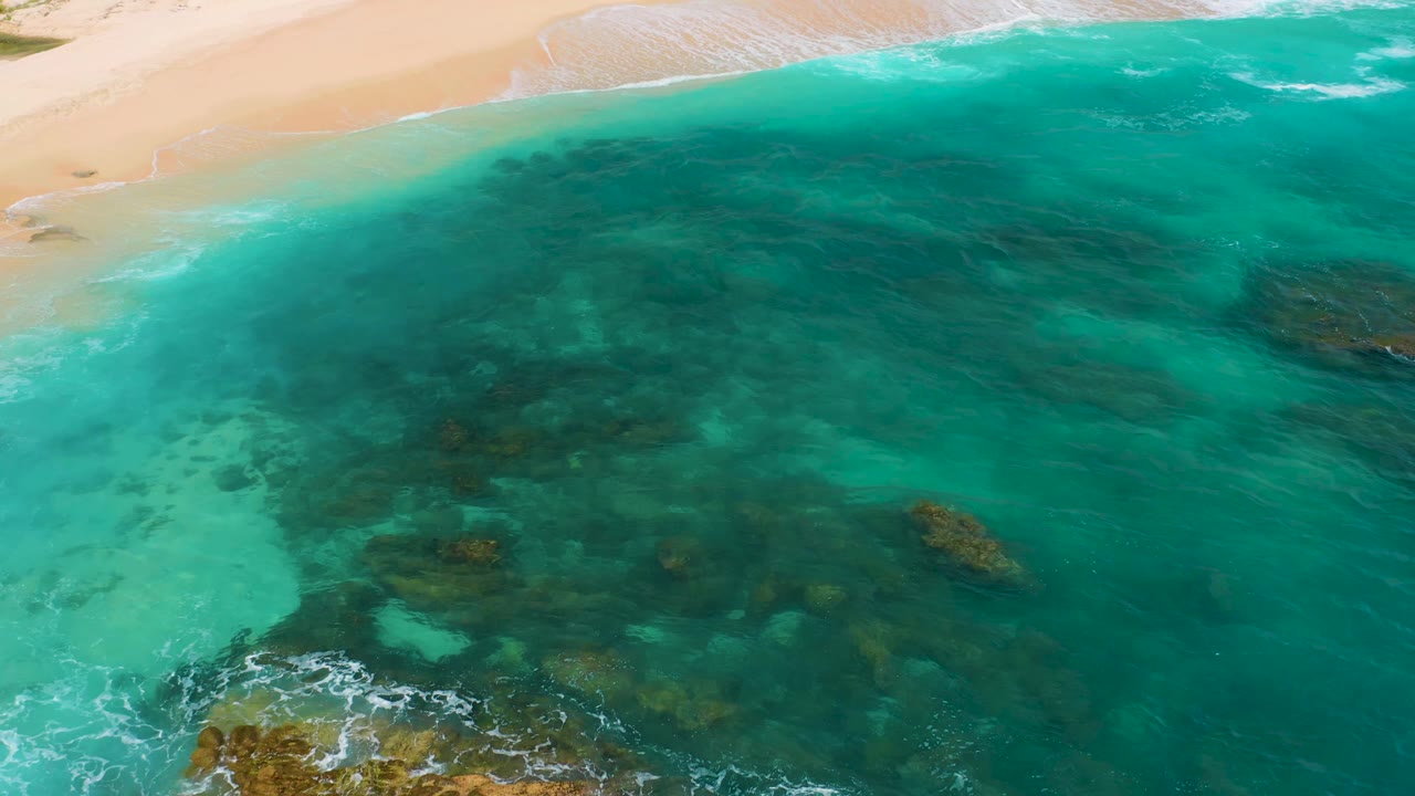 Aerial frame at Secrets Marquis Los Cabos, Mexico, with a wider band of green-blue water, scattered dark rock patches below the surface, sand along the top edge, and no visible people.