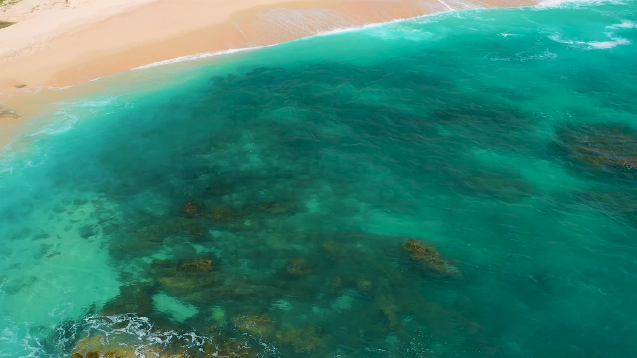 Aerial view at Secrets Marquis Los Cabos, Mexico, showing turquoise ocean filling most of the frame, darker submerged rocks across the lower half, white surf near the sand, and no visible people.