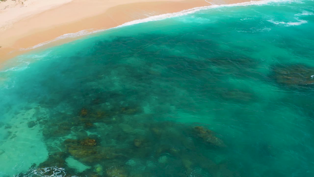 Aerial frame at Secrets Marquis Los Cabos, Mexico, with pale sand running across the top, a soft white wave line, broad turquoise water, darker underwater patches, and no visible people.
