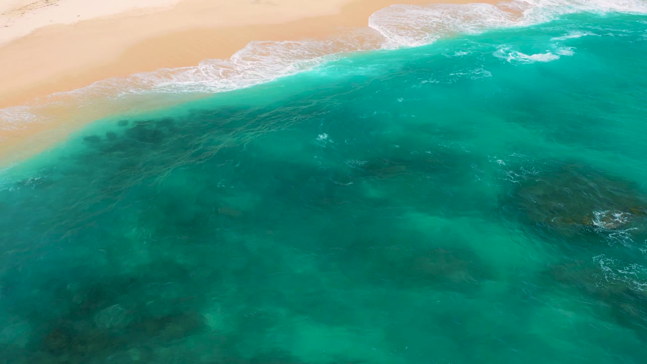 High overhead view at Secrets Marquis Los Cabos, Mexico, showing pale sand, a bright surf band, broad turquoise water, scattered darker patches, and no visible people.