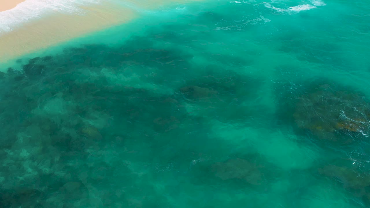 Aerial frame over Secrets Marquis Los Cabos, Mexico, with green-blue water, darker rock patches beneath the surface, soft wave texture near the sand, and no visible people.