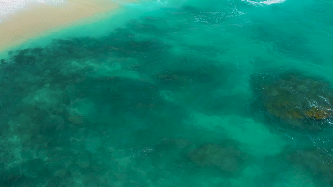Top-down coastal water view at Secrets Marquis Los Cabos, Mexico, where turquoise shallows reveal broad dark rock forms below the surface and a pale sandy edge remains at the top, with no visible people.