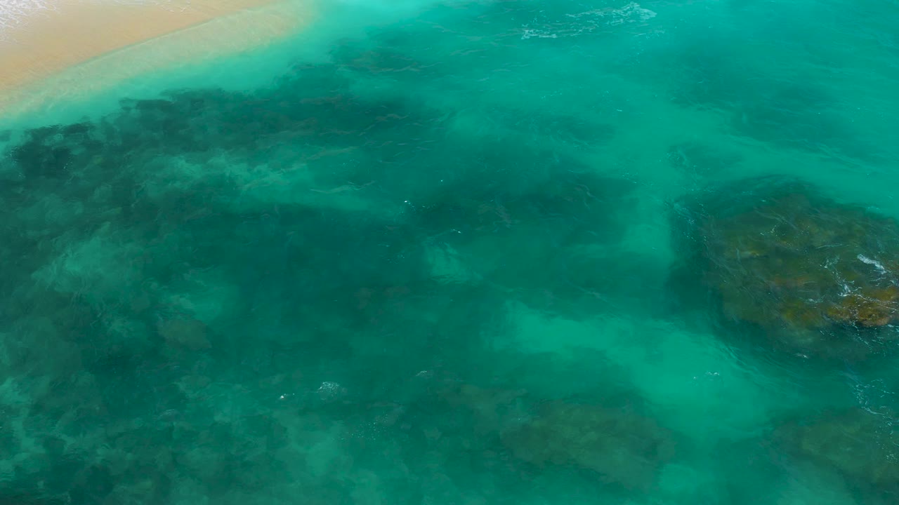 Steep aerial frame from Secrets Marquis Los Cabos, Mexico, showing a larger field of green water, muted underwater rock masses, and a sliver of sandy shoreline at the upper left, with no visible people.
