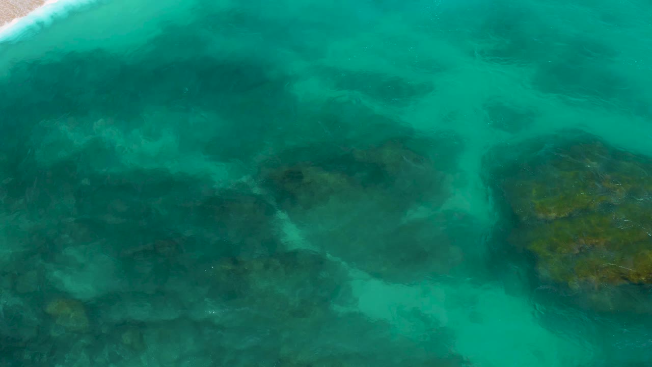High overhead frame from Secrets Marquis Los Cabos with translucent aqua water crossing darker underwater shapes and a narrow band of white surf at the upper left, with no visible people.