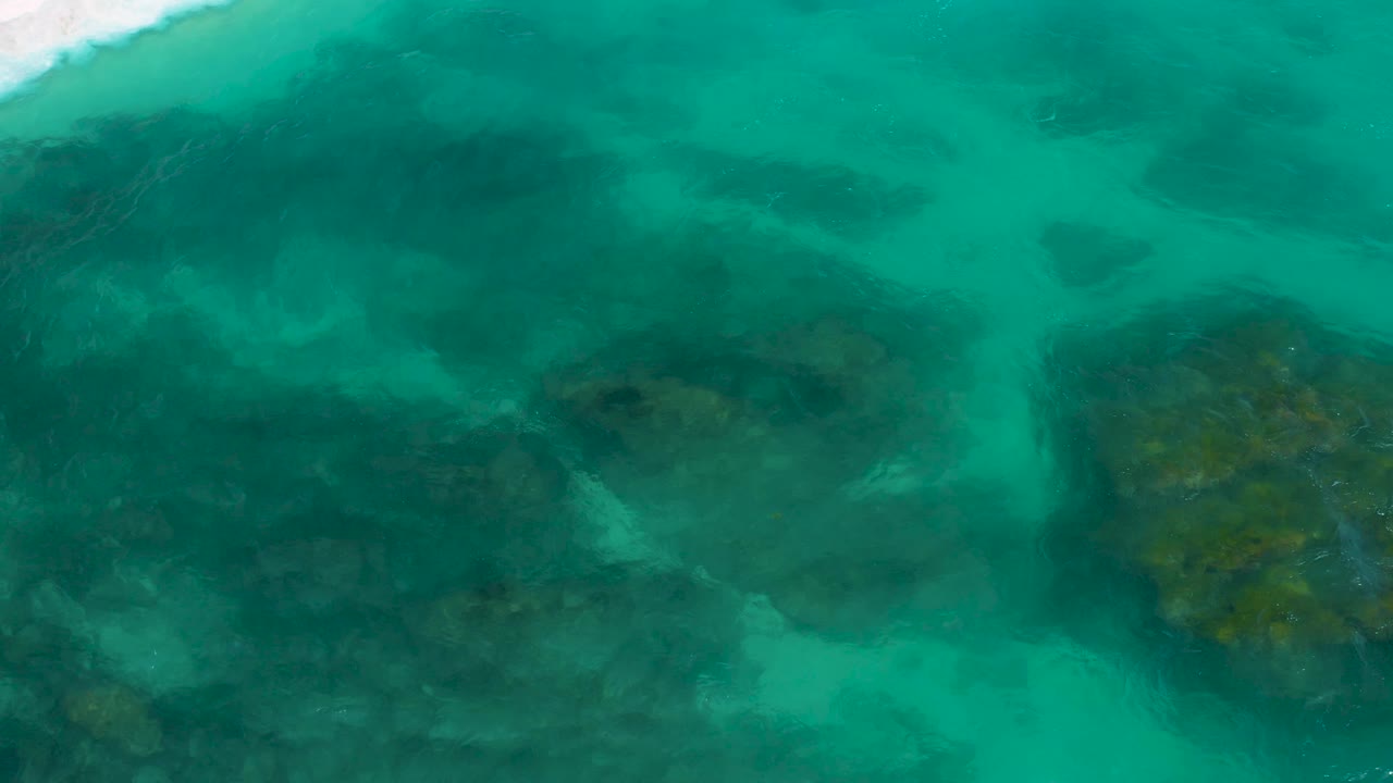 Overhead Secrets Marquis Los Cabos water frame with a brighter white surf band along the upper left, mottled blue-green water, dark underwater patches, and no visible people.