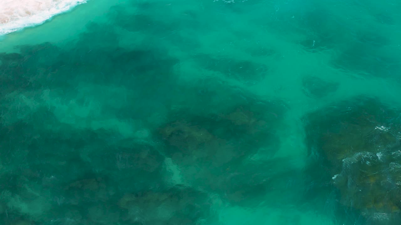 Secrets Marquis Los Cabos overhead frame showing clear aqua ocean water spread across the frame, darker reef-like formations below the surface, a partial surf edge, and no visible people.