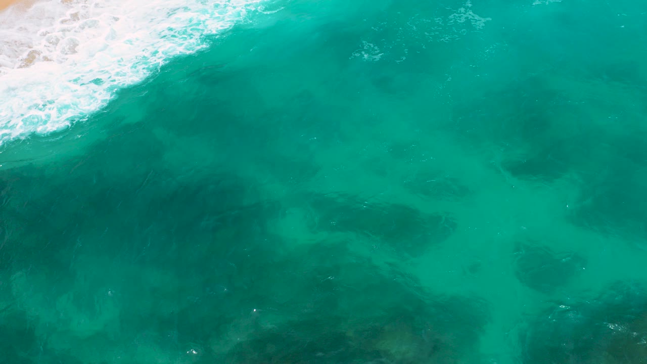 Top-down view of turquoise water and dark submerged patches near a sandy shore at Secrets Marquis Los Cabos, Mexico, with white surf entering from the upper left and no visible people.