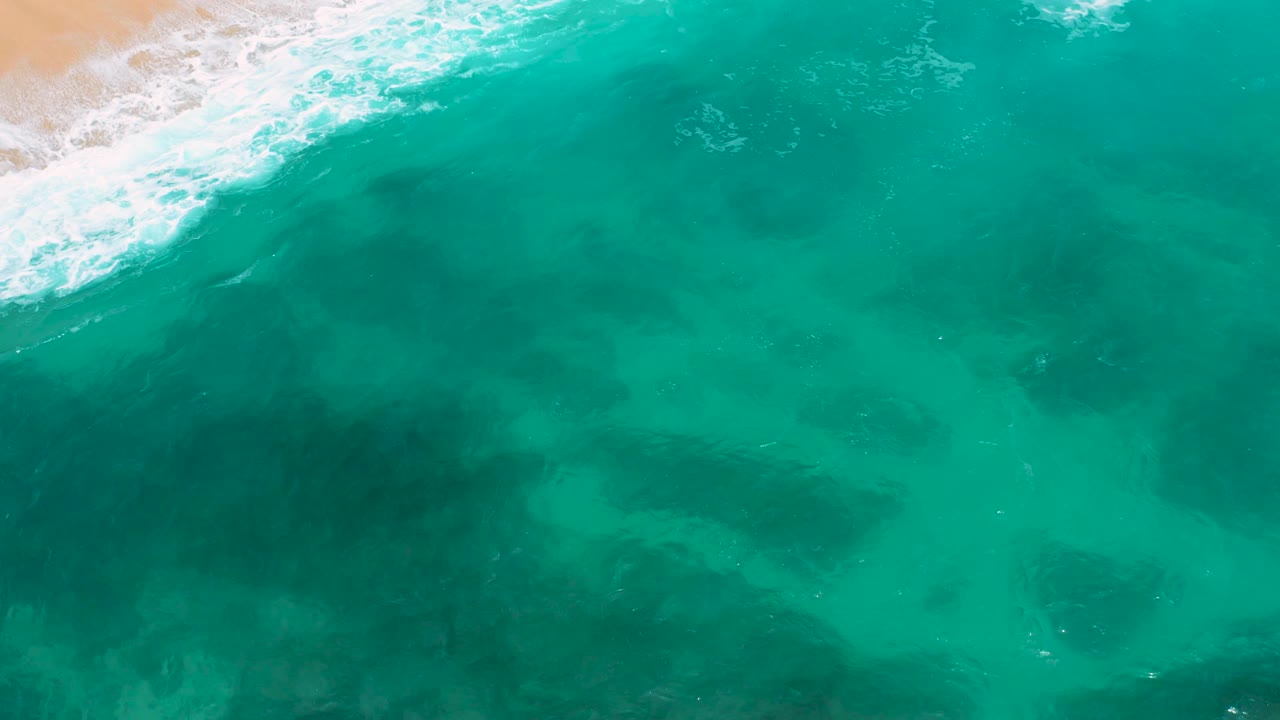 Steep aerial frame of turquoise ocean at Secrets Marquis Los Cabos, Mexico, with a narrow tan sand strip in the upper left, foamy surf along the edge, and no visible people.