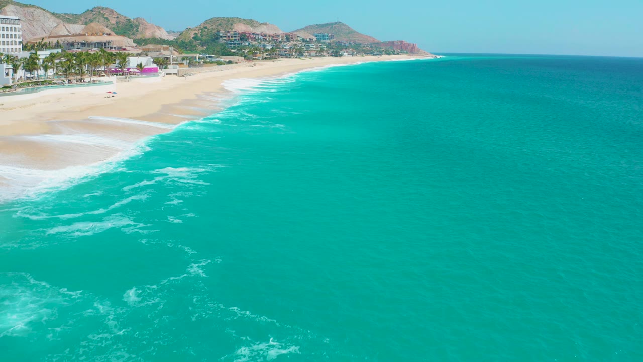 Aerial view at Secrets Marquis Los Cabos, Mexico, looking along the sandy beach as foamy surf traces the shore and open turquoise water fills the lower and right side, with no visible people.