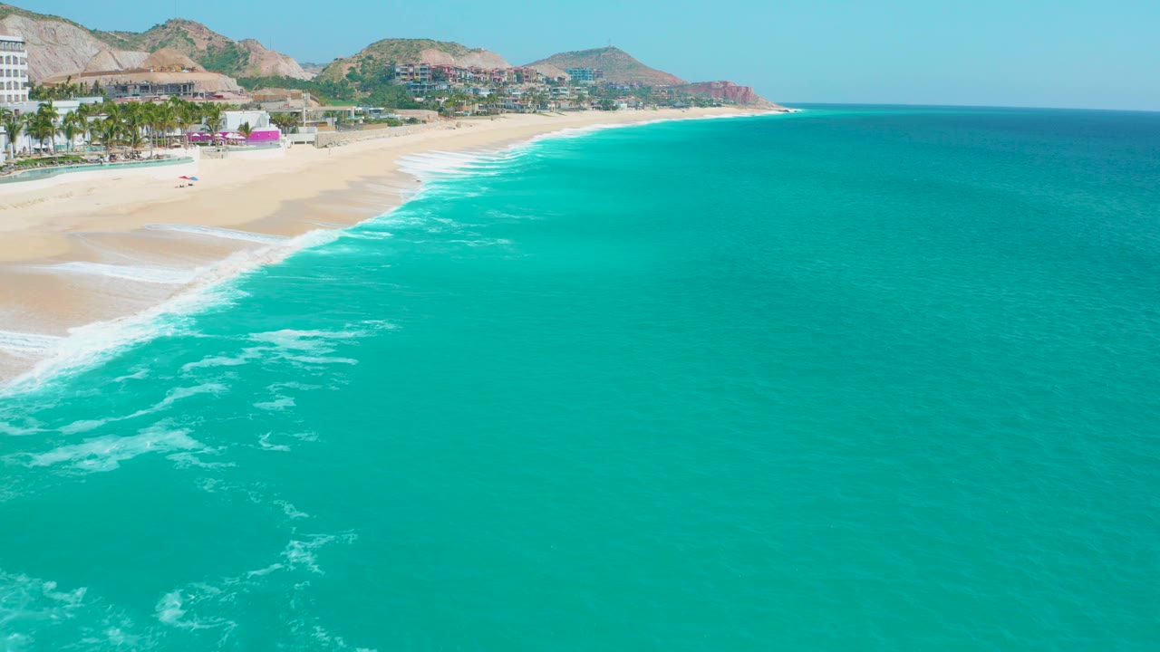 Aerial frame over Secrets Marquis Los Cabos, Mexico, with the beach and resort strip compressed along the left edge, a broad expanse of turquoise sea to the right, distant hills, and no visible people.