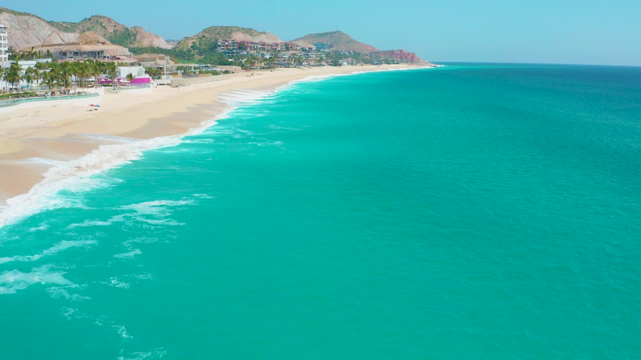 Secrets Marquis Los Cabos, Mexico coastline seen from above, with pale sand at left, bright white surf breaking in the midground, resort buildings and palms behind it, hills beyond, and no visible people.