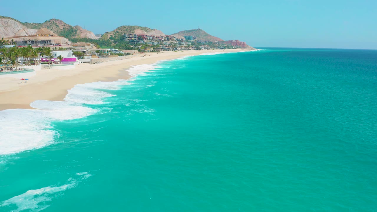 Aerial frame from Secrets Marquis Los Cabos, Mexico, showing a broad sweep of blue-green ocean, foamy waves near the left beach edge, hillside terrain, shoreline buildings, and no visible people.