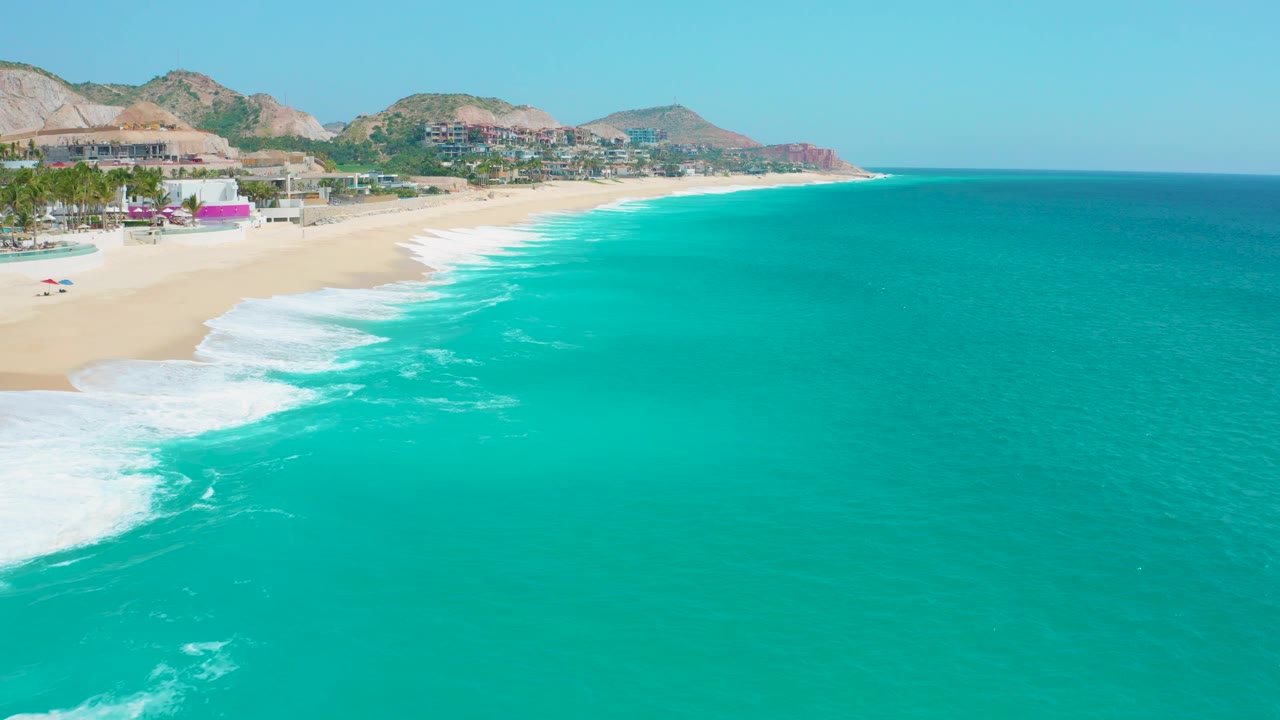 Aerial shoreline view at Secrets Marquis Los Cabos, Mexico, where the turquoise water fills the right side, white surf marks the beach curve, hills sit behind the buildings, and no visible people appear.