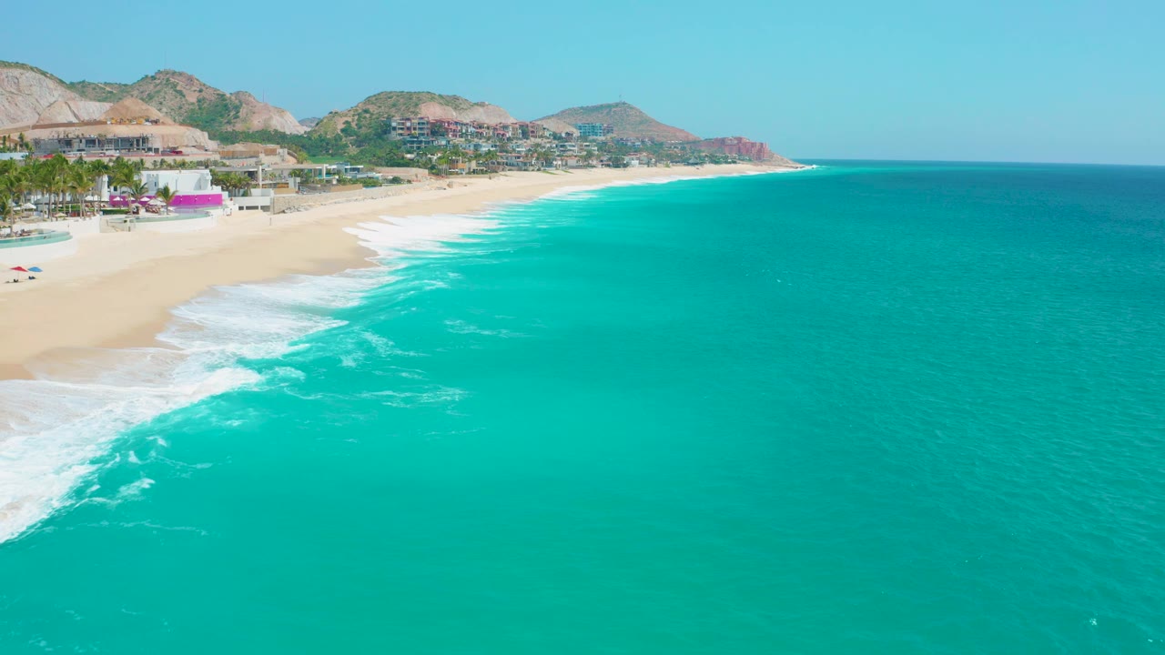 At Secrets Marquis Los Cabos, Mexico, the frame shows bright turquoise water across most of the view, surf spreading along the sandy shore, hillside development in the background, and no visible people.