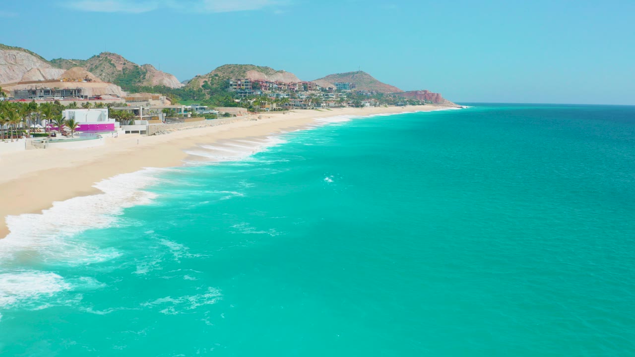 Opening aerial view of Secrets Marquis Los Cabos, Mexico with turquoise ocean in the foreground, a curving sandy beach, low resort buildings, palms, hills, and tiny beach figures visible.