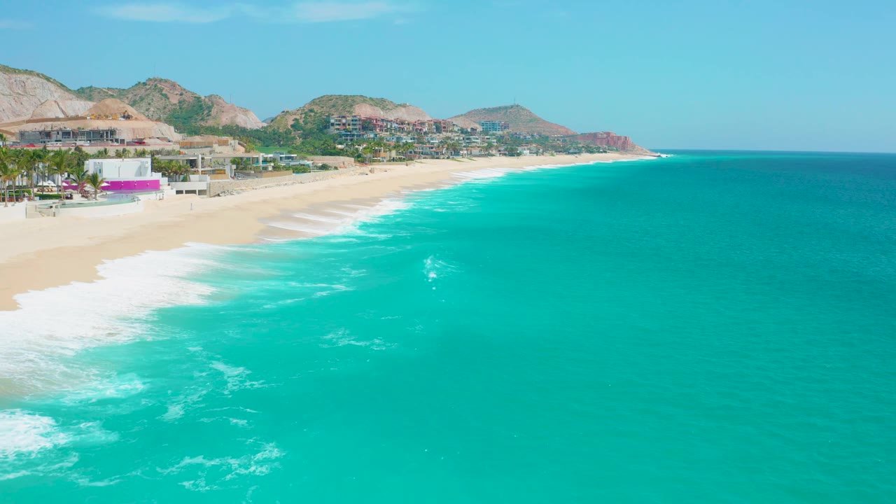Aerial view at Secrets Marquis Los Cabos, Mexico with more open turquoise water dominating the right side, surf rolling onto sand at left, resort buildings behind the beach, and distant hills under clear sky.