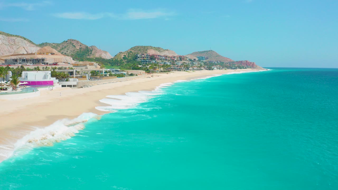 At 0:00, an elevated view over Secrets Marquis Los Cabos, Mexico shows turquoise water, white surf, an empty sand beach, hillside buildings, and low mountains with no visible people.
