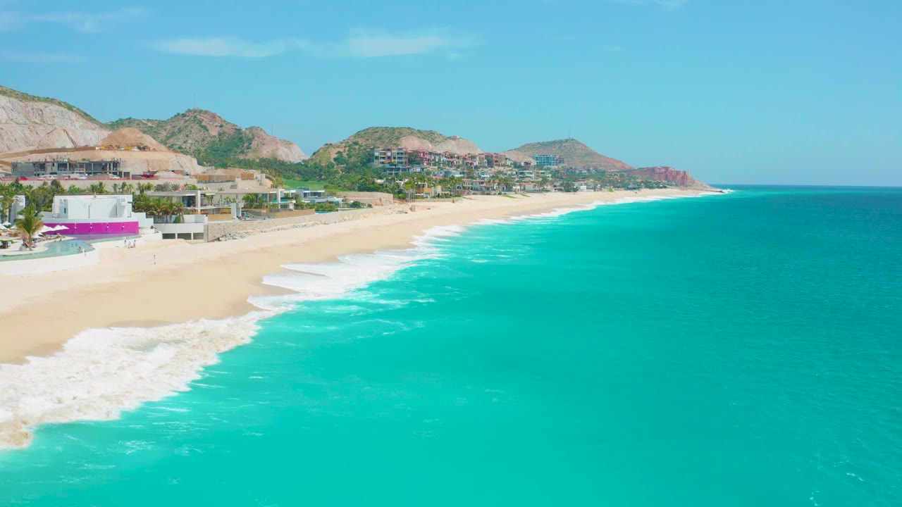 At 0:02, turquoise water fills most of the frame as white waves reach the empty beach below hillside development and distant mountains in Secrets Marquis Los Cabos, Mexico.