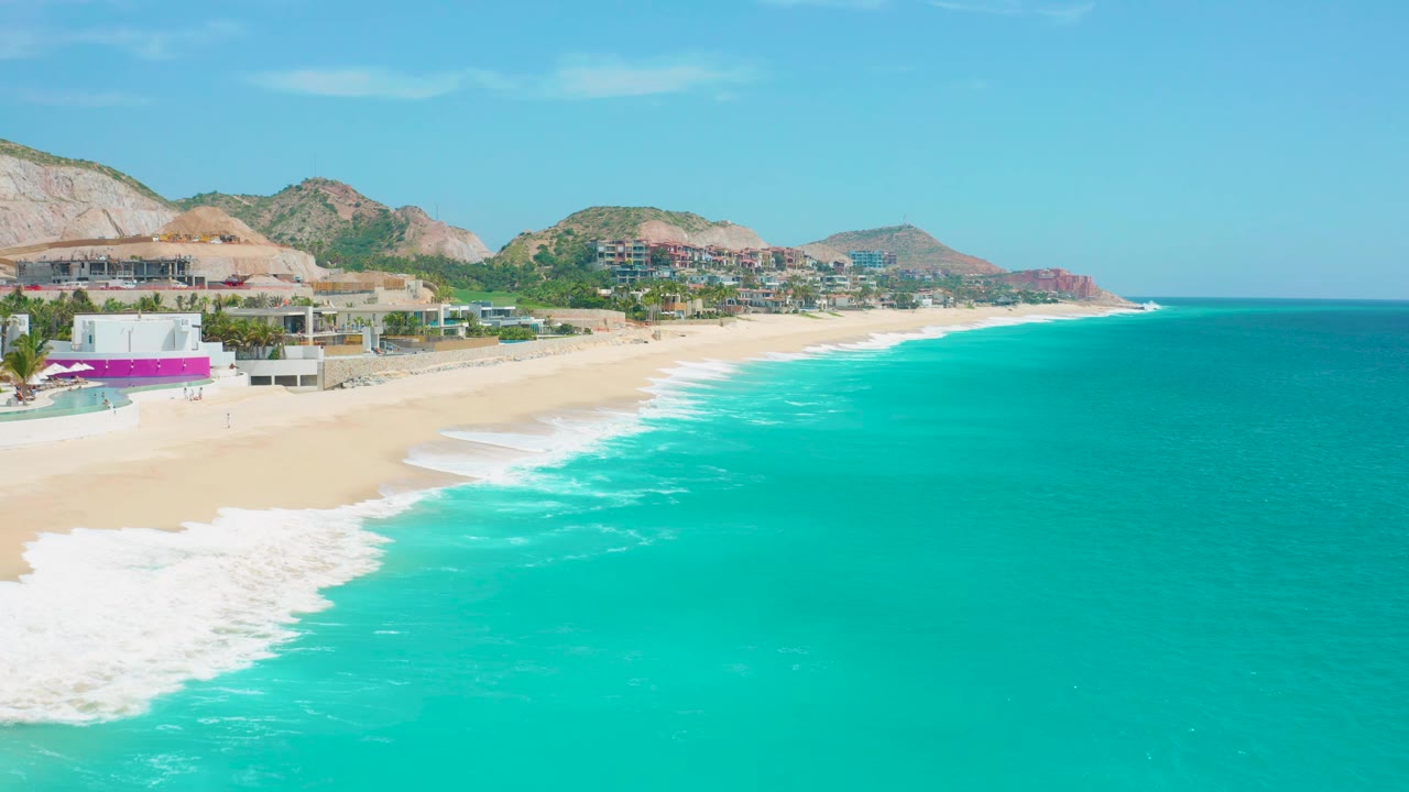 Foamy surf reaches a sandy beach beneath coastal buildings and rocky hills.