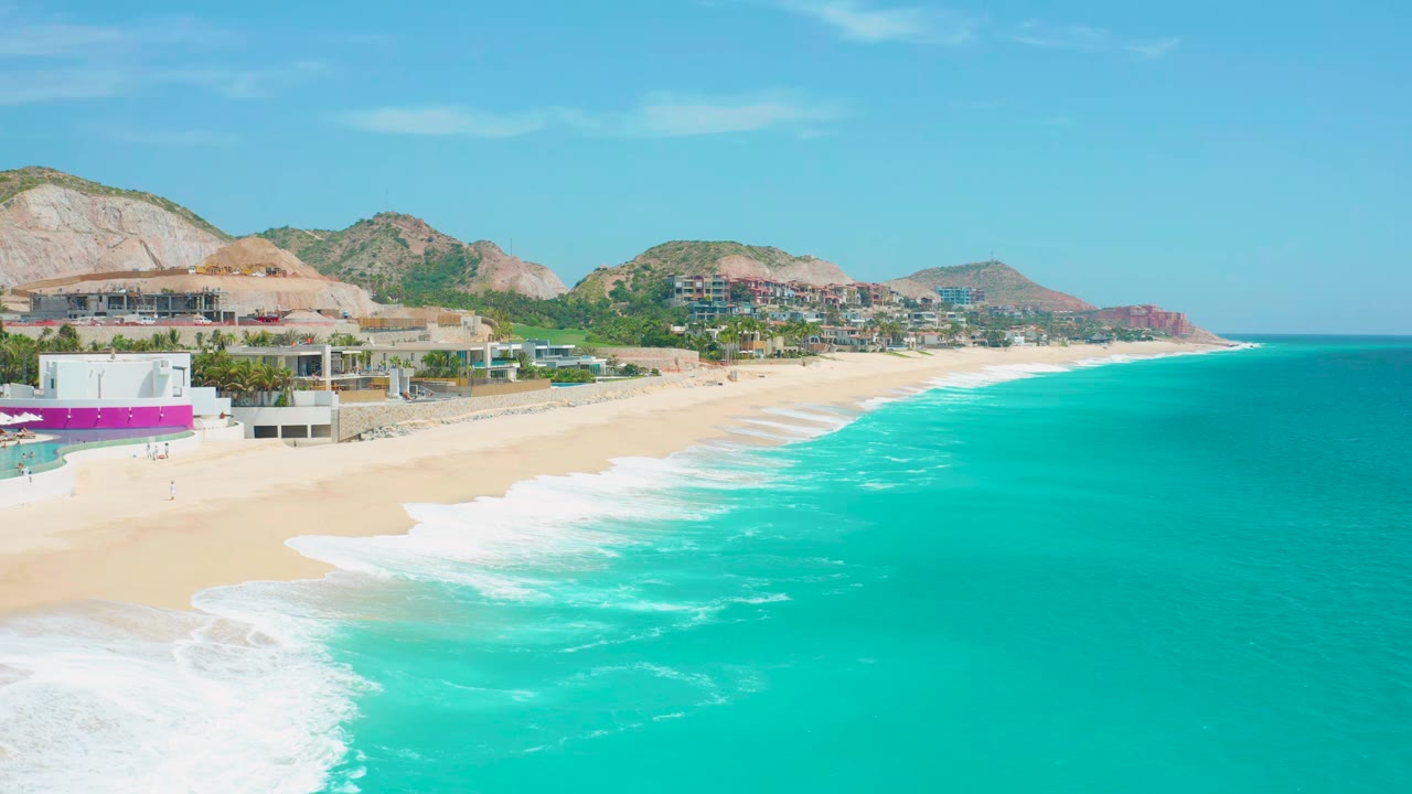 Aerial view at Secrets Marquis Los Cabos, Mexico, showing a longer band of white surf along the sandy beach, low resort buildings at left, hills in back, and no visible people.