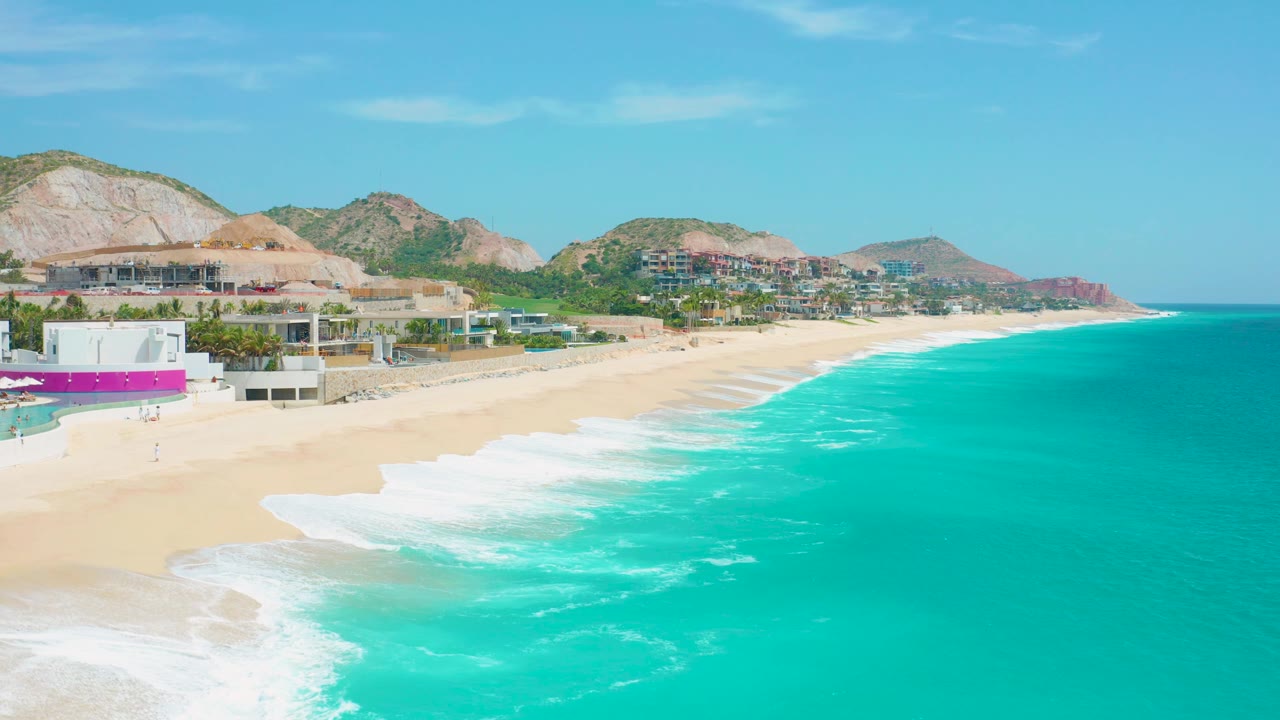 Bright aerial frame at Secrets Marquis Los Cabos, Mexico, with aqua water spreading across the right side, foam reaching the sand, beachfront buildings at left, hills beyond, and no visible people.