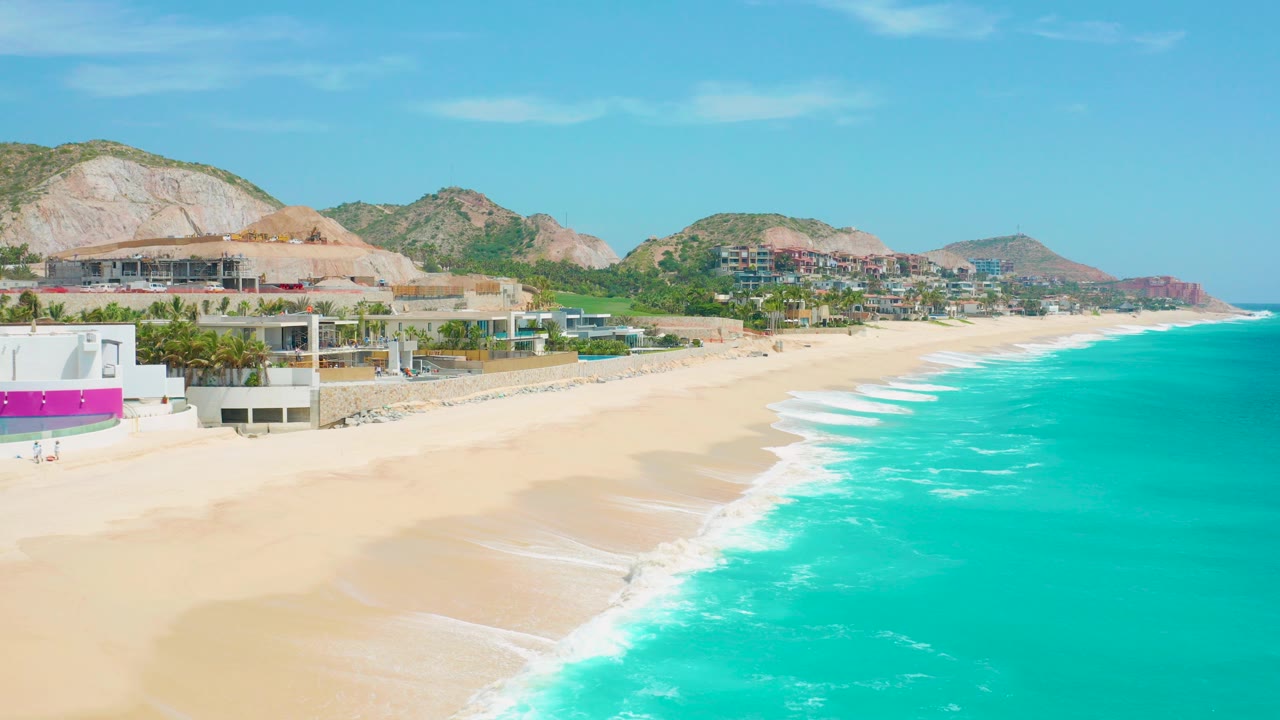 Opening aerial frame over Secrets Marquis Los Cabos in Mexico, with turquoise water along the right, pale sand below, resort buildings and terraces at left, rocky hills behind, and tiny beachgoers with no readable faces.