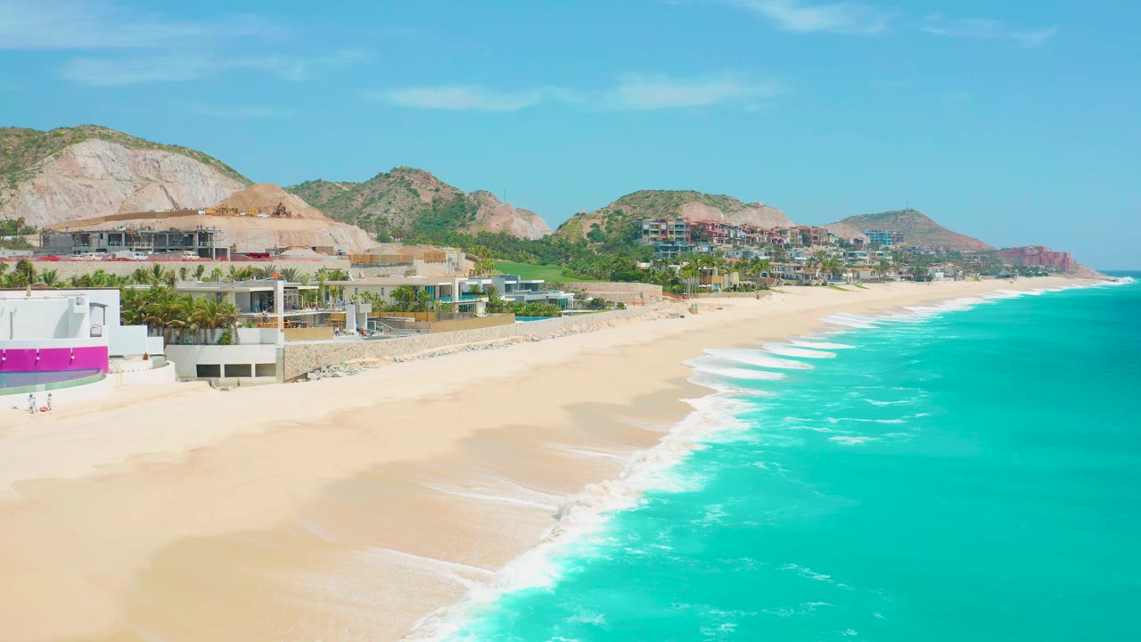 Wide coastal view with turquoise water, sand, terraces, palms, and hillside buildings.