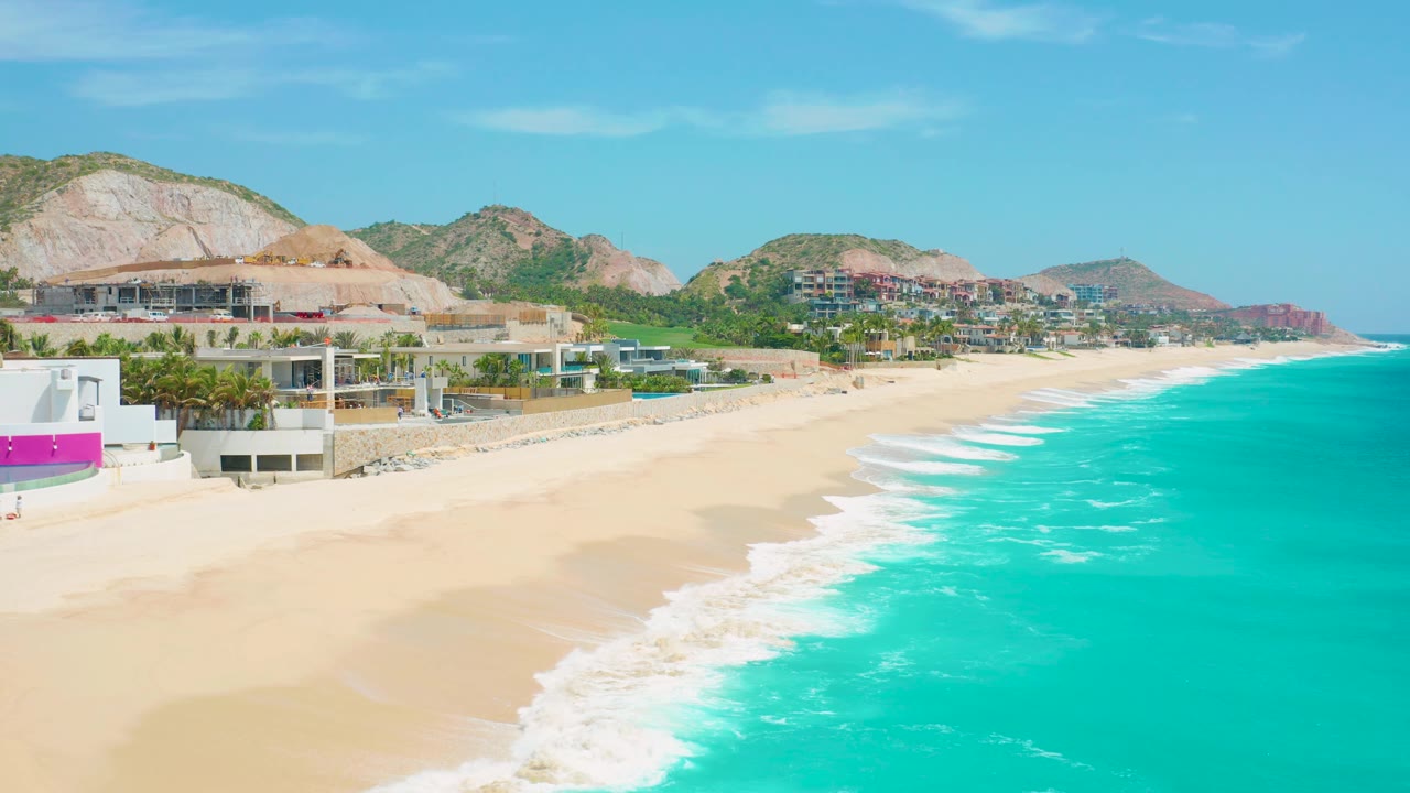 Third aerial frame over Secrets Marquis Los Cabos in Mexico, with a stronger band of white surf at the water edge, broad empty-looking sand, low resort structures, palms, and dry hills beyond.