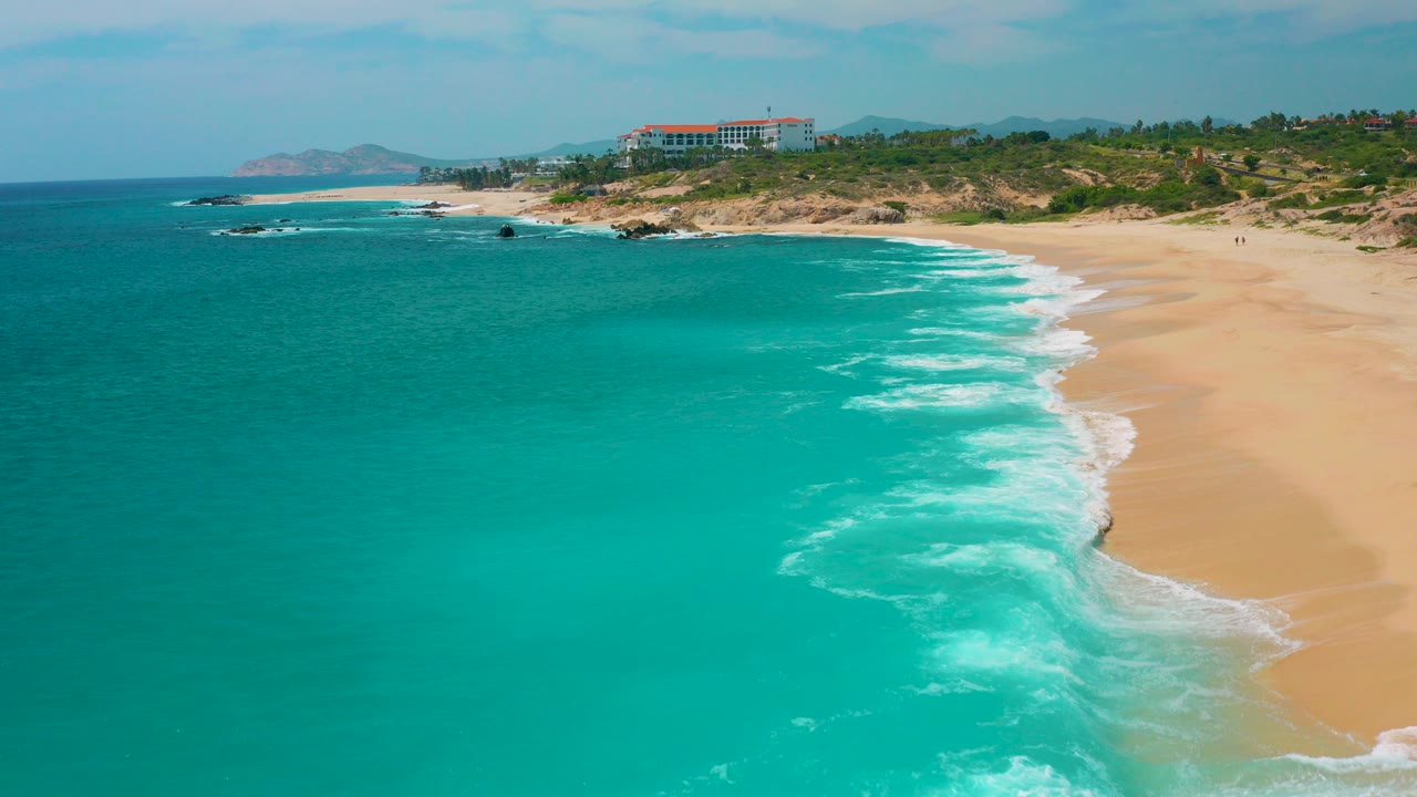 Aerial frame at Secrets Marquis Los Cabos, Mexico, showing turquoise water at left, white surf on a wide sand beach, a low green ridge, a distant red-roofed building, and no visible people.